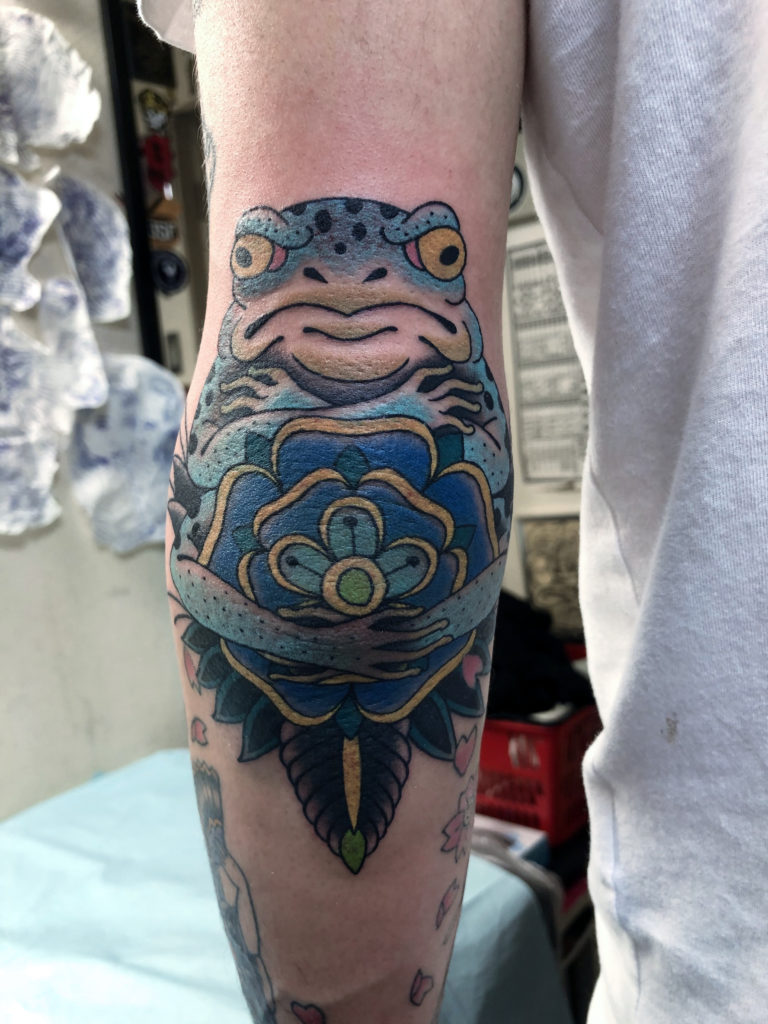 花の後ろで腕組みをするカエルが描かれたタトゥー